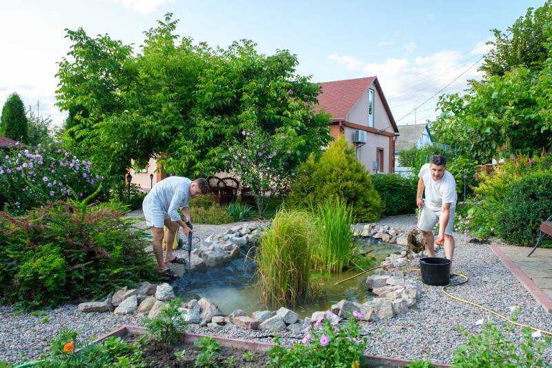Mature father and young adult son cleaning artificial garden pond bottom with high-pressure washer nozzle from
